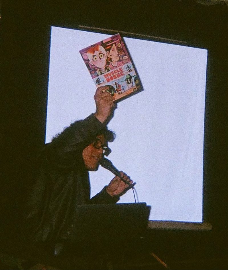Nate Garcia, a man with light brown skin, eyebrow length bangs and shoulder-length brown hair, wearing black, thick framed glasses and a leather jacket, standing in front of a blank projector screen in a dark room, holding a mic to his mouth with his left hand, and holding up a copy of his comic book, "Muscle Horse," with his right hand.