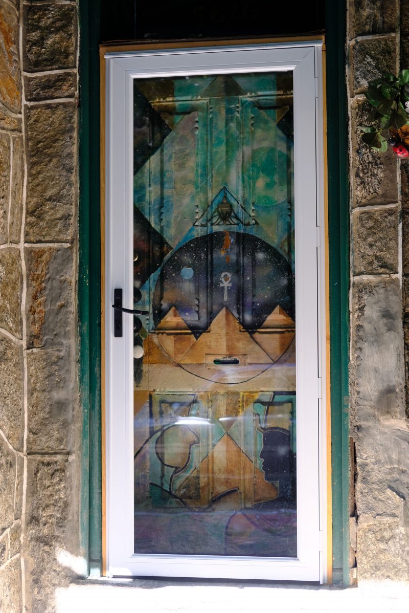 Front door of the Sun Ra house, painted on which are egyptian symbols and figures, stylized with geometric shapes with textured colorful patterns within them. Edges of the door are partially obscured by a closed glass door with a white frame. The exterior entranceway to the door is made of stone.