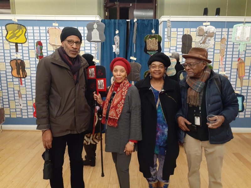 Four African American artists inside at a gallery opening pose for a picture, looking happy, with art pictured in the background.