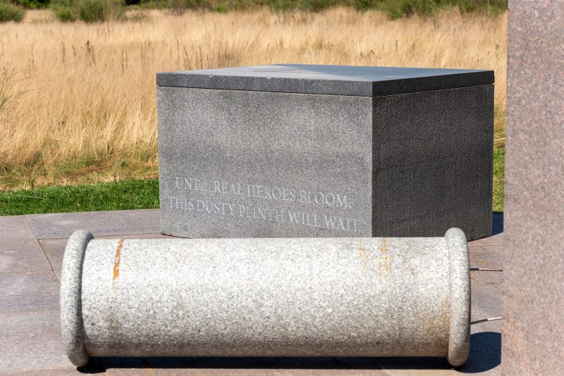 Detail shot of a public artwork made out of stone with various tone objects sitting on a stone platform in a gray field. In the front, a cylindrical object resembling a large mortar; in the back a rectangular stone structure with the words inscribed: "Until real heroes bloom, this dusty plinth will wait." 