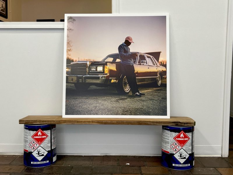 Large photograph of a Black man in a pink har, sweater, black pants and black shoes, leading up against a vintage car with the headlights on and the trunk popped open, taken as the sun sets; the photo leans against the gallery wall and sits on a wooden plank that's propped up by two large cans of gasoline sitting on the gallery floor.