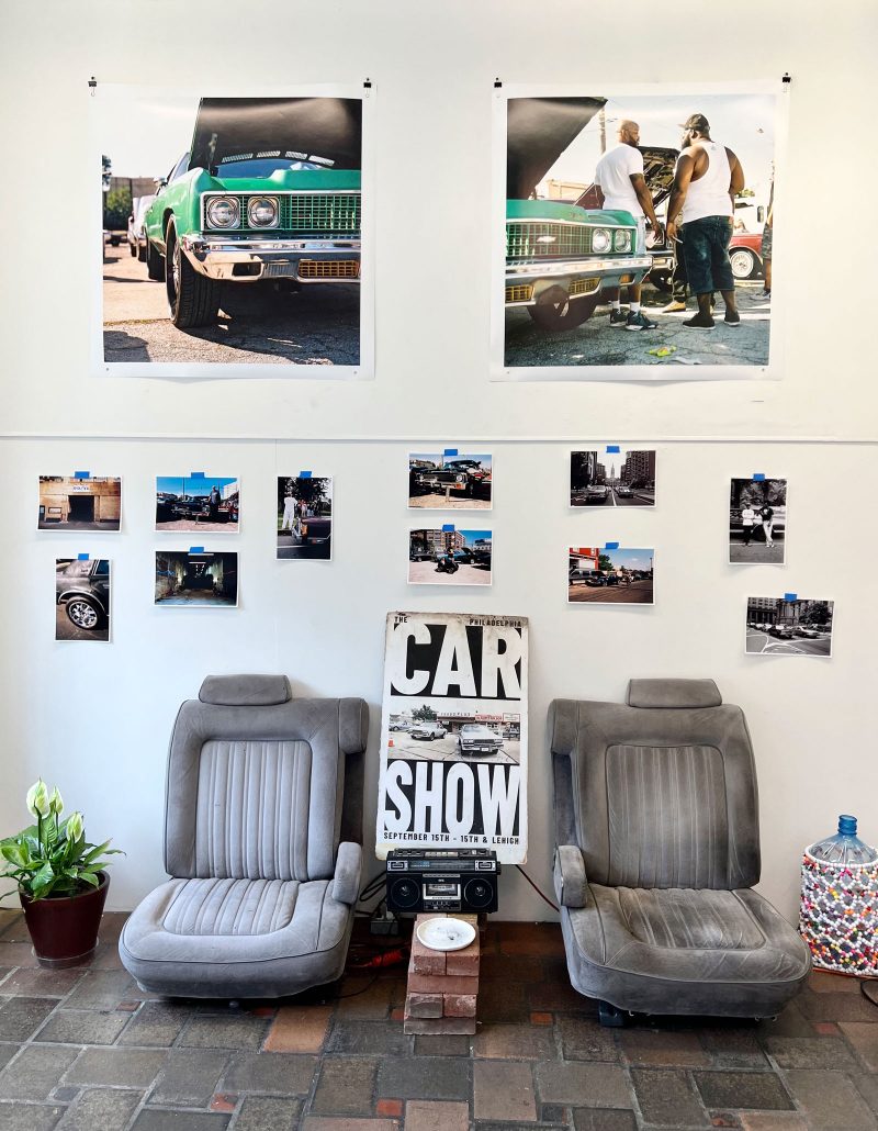 Installation view of two large photos of cars and people working on them; 11 smaller photos of cars, lots, people, and streets, below it; and on the floor below all of the pictures, two car seats between which are bricks with a boombox and a sign that says "CAR SHOW" leaning on it.