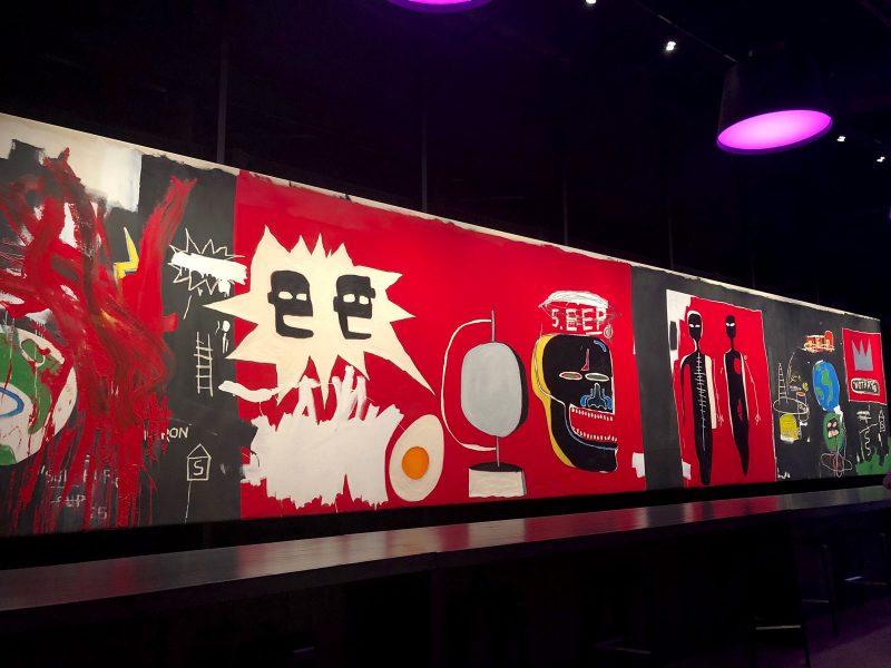 Installation view of a large abstract painting on a gallery wall with a mainly red and black background, with abstract figures painted in black and other images such as a globe, crown, egg and traces of paint over other areas.