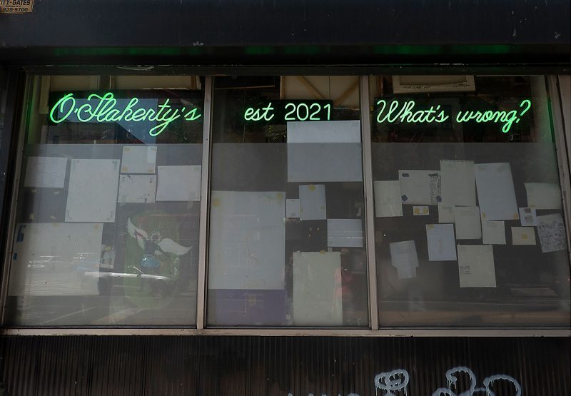Exterior view of O'Flaherty's gallery, which has "O'Flaherty's," "est 2021," and "What's wrong?" in neon signs hanging in 3 windows, through which you can see multiple artworks taped to the windows.