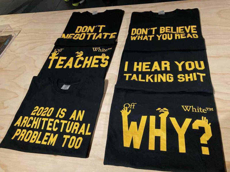Laid out in two straight rows on an unpainted plywood table, six neatly-folded, black t-shirts feature yellow text and designs that spell out political poster-like statements and a question, including “Don’t Believe What You Read” and “2020 is an Architectural Problem Too.”