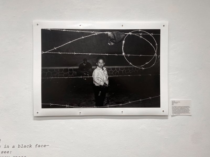 A black and white photo, pinned to the wall of a gallery. It showing a boy of color, standing behind barbed wire at night.