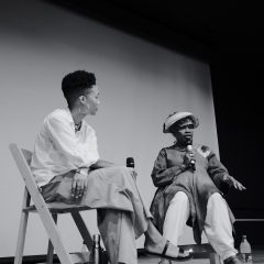 In a black and white photo, two Black women sit on folding chairs on a stage facing an audience that is not seen, and both hold microphones in their hands, while one of them speaks.