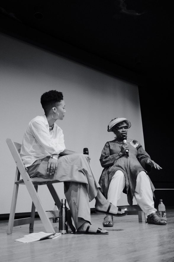 In a black and white photo, two Black women sit on folding chairs on a stage facing an audience that is not seen, and both hold microphones in their hands, while one of them speaks.