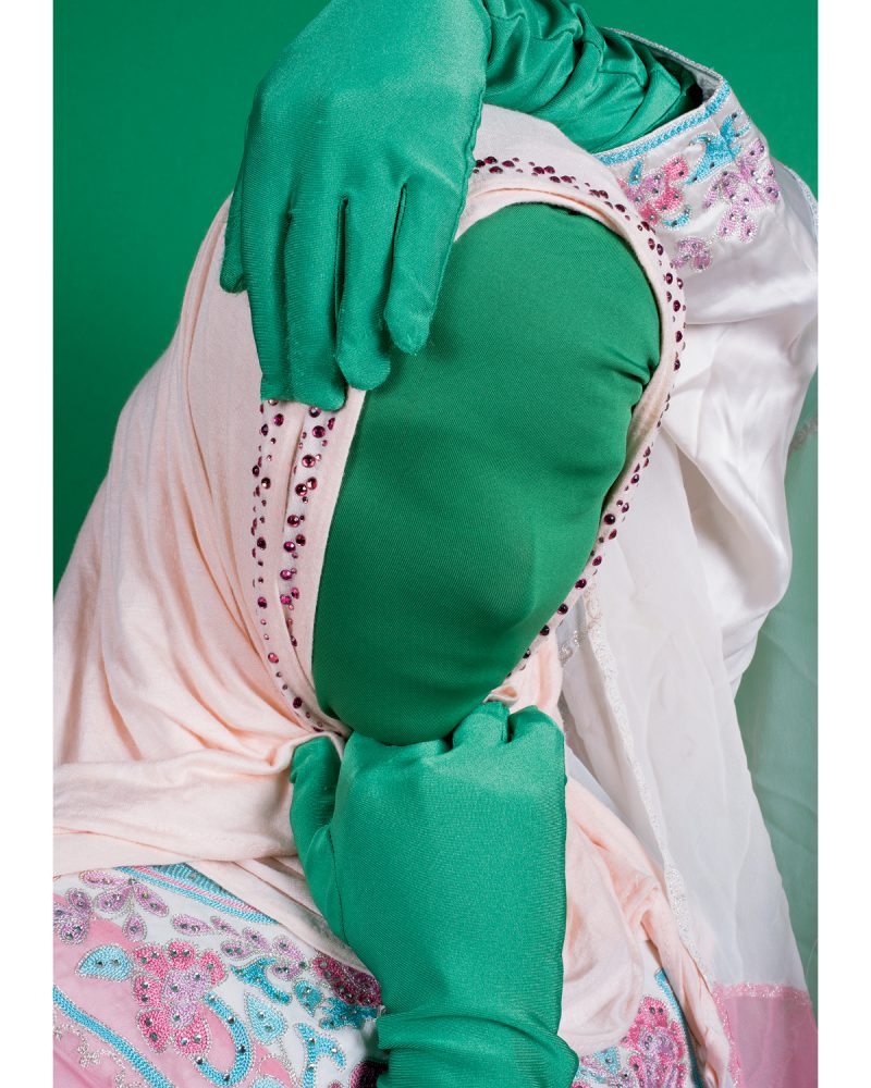 A color photo shows a woman in closeup with her hands touching her head and chin. She is covered in fabric, with a pink scarf and tunic and green gloves and a green cloth over her face. The background is green.