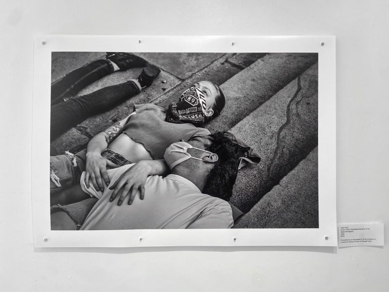 A black and white photo pinned to a gallery wall.  It depicts two people wearing masks, lying together on the ground.