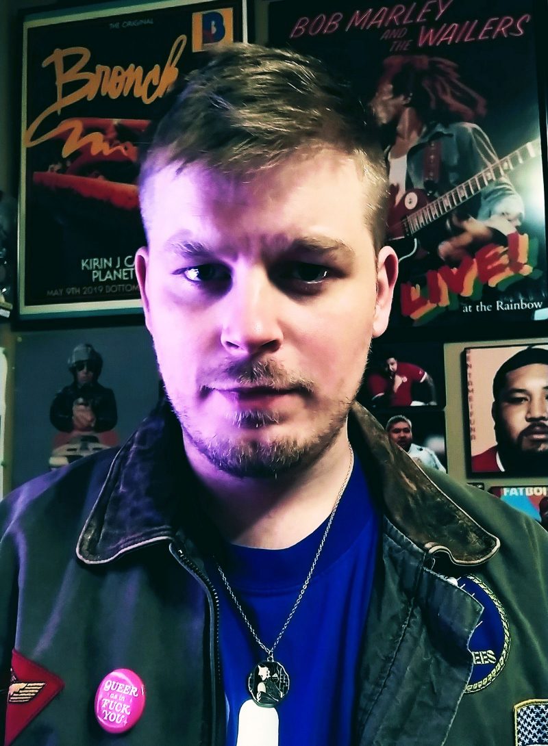 James Dillenbeck, a white man with short blonder hair and a dirty blonde mustache, soul patch, and short beard, wearing a green jacket with a pink pin that says "queer as in fuck you," and a medallion necklace, looking straight into the camera with a series expression; half of his face is lit with a purple light.