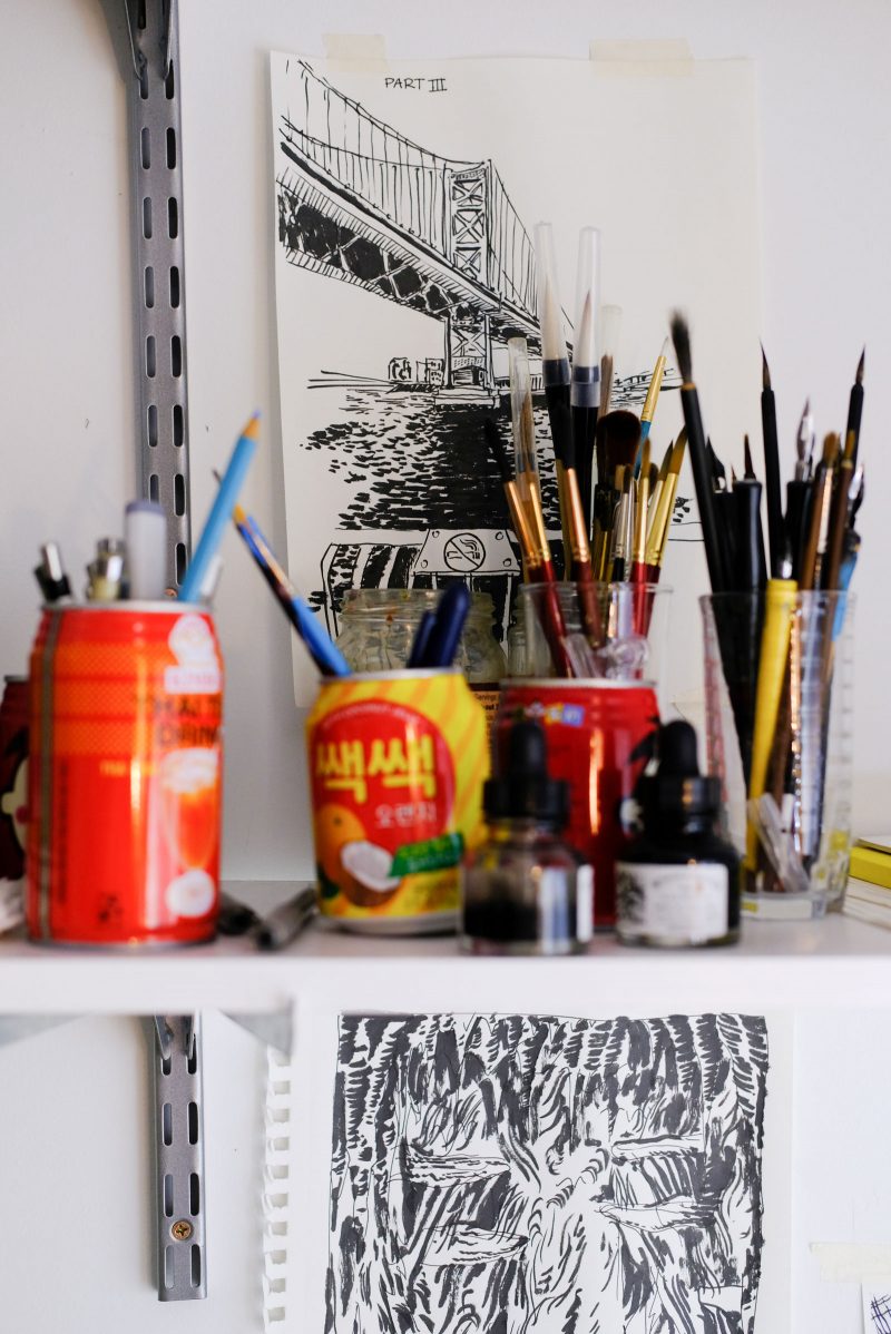 A color photo shows a white shelf that holds bottles of ink and cans and glasses filled with pencils and brushes. On the wall behind is a black ink drawing of the Ben Franklin bridge, and below the shelf another black ink drawing of vegetation.