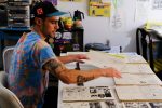 A color photo shows a man wearing a black baseball hat with a blue brim, and a bright blue, red and yellow tie dye t-shirt sitting at a table assembling some papers for a comic book.
