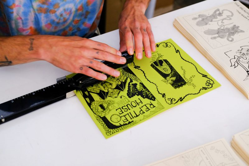 Barevná fotografie ukazuje mužské potetované ruce a paže a plátek tohoto pestrobarevného trička s kravatou, když sešívá obálku a stránky komiksu černou průmyslovou sešívačkou.