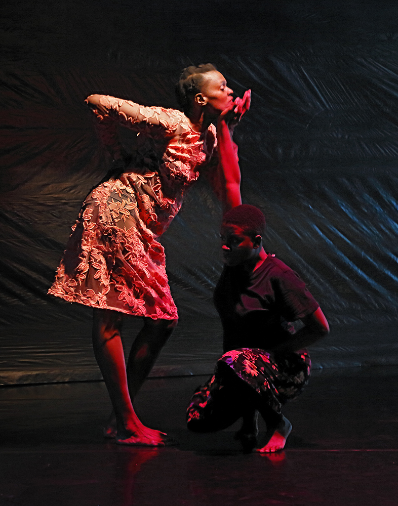 A color photo shows two Black women dancers in a space with a black floor and black plastic backdrop. 