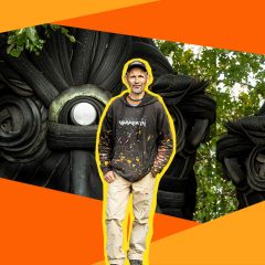 Pedro Ospina, a Colombian man with short salt and pepper hair, wearing a paint covered black hoodie, tan paints, a black shirt, and work boots, standing in front of a massive sculpture of a face, made out of tires, which is inside of his outdoor sculpture garden.