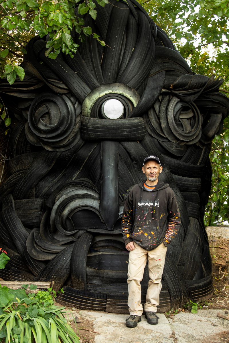 Pedro Ospina, a Colombian man with short salt and pepper hair, wearing a paint covered black hoodie, tan paints, a black shirt, and work boots, standing in front of a massive sculpture of a face, made out of tires, which is inside of his outdoor sculpture garden.