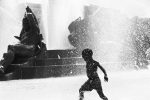 A black and white photo shows a young Black boy running through a large fountain with sculptural figures in the background.