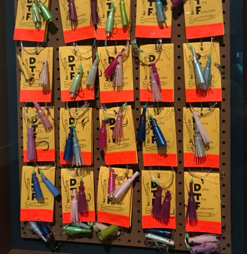 On a peg board, rows of brightly colored paper and plastic. Each item has two flared plastic pieces connected by fishing line, attached to paper cards that read DTF (Duke the Fisherman).
