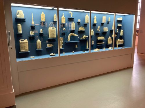 Inside a large museum display case, there is an array of bleached plastics that have been decoratively painted with black ink.