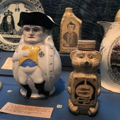 Installation view at a museum. A honey bear has been decoratively painted, and behind it a jug has been illustrated in a similar manner. Surrounding these artworks are several portraits on ceramics.