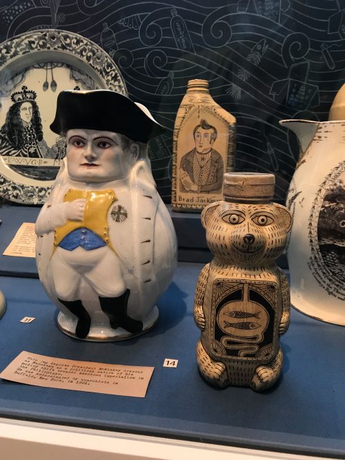 Installation view at a museum. A honey bear has been decoratively painted, and behind it a jug has been illustrated in a similar manner. Surrounding these artworks are several portraits on ceramics.