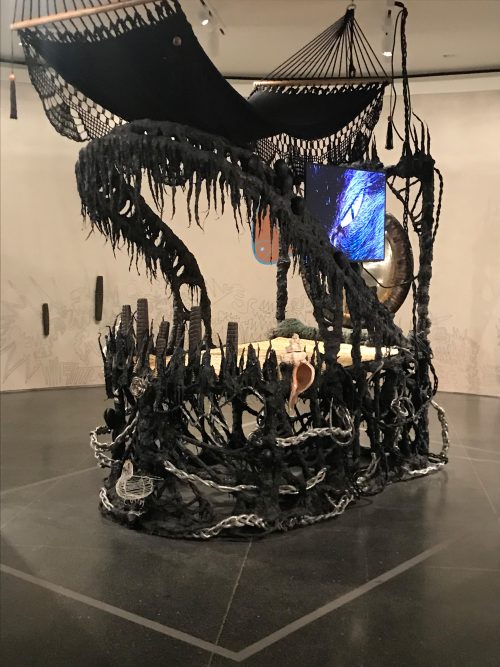 In a museum gallery, a black mixed media sculpture resembles a dragon. The sculpture is built around a mattress and gong, and contains many intricate details.