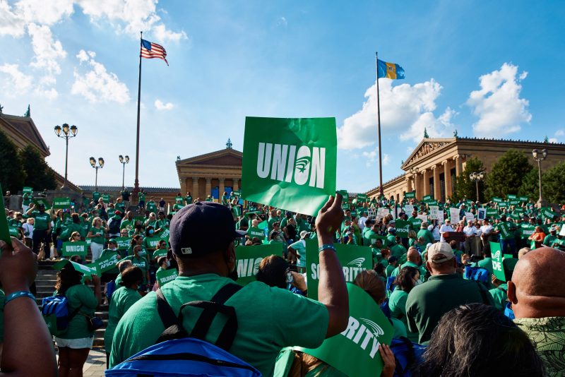 Standing in front of the Philadelphia Museum of Art, a large rally of protestors are gathered wearing green shirts and blue backpacks. Most are holding green protest signs saying "union," "solidarity," or "AFSCME."