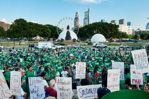 A large rally of protestors is gathered, most wearing green shirts and holding protest signs saying "union," "solidarity," or "AFSCME." In the foreground, handwritten protest signs stand out. In the background is the Washington Monument Fountain, a Ferris Wheel, and the Philadelphia skyline