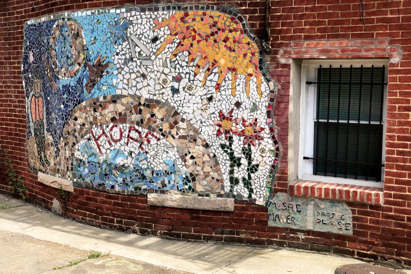 A photo shows a red brick wall of a building with a window with bars protecting it and a colorful ceramic tile mural of hope. 