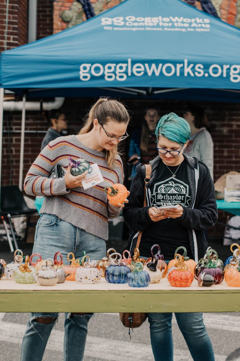 A colorful photo shows two young women looking at small glass pumpkins sitting on a table outside at what looks like an art fair. In the background a tent announces “Goggleworks.org”