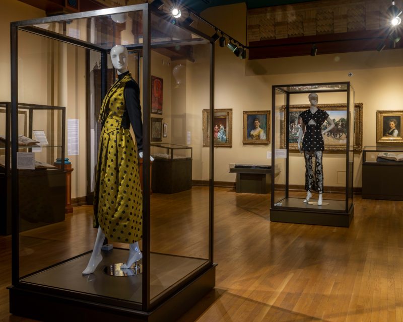 Two mannequins wearing decoratively patterned clothing inside a museum stand surrounded by large books in display cases and traditional style oil paintings on the wall.