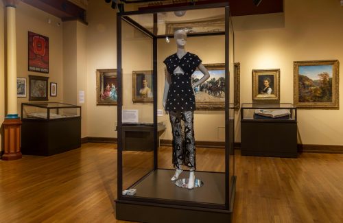 In a museum gallery, a mannequin wearing a patterned mini dress with cutout details and a lace skirt stands in front of oil paintings, posters, and large books in display cases.