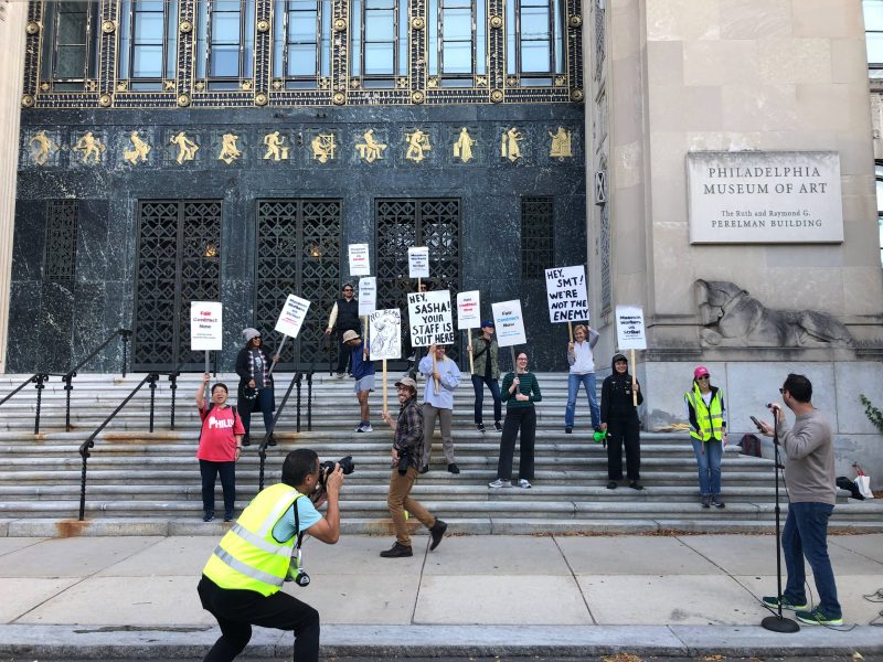 A group of striking Philadelphia Museum of Art Union workers hold hand-made and printed picket signs outside of the museum's Perelman Building.