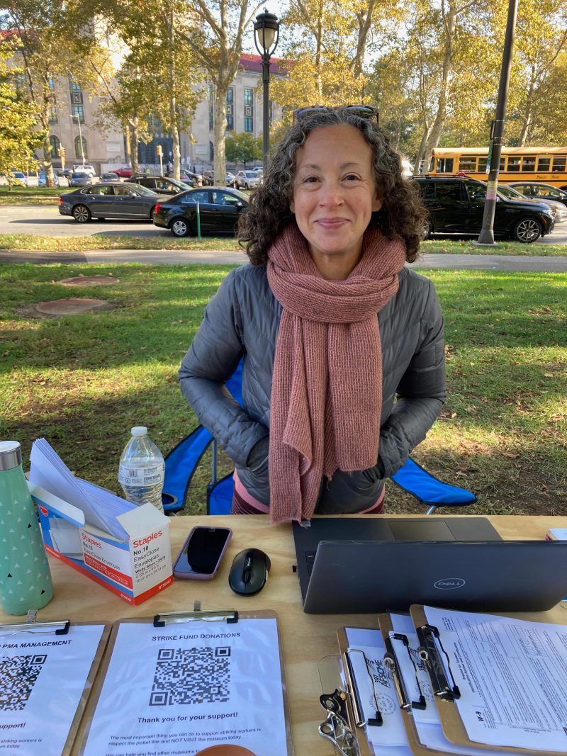 A woman wearing a black jacket and mauve scarf sits outside the the Philadelphia Museum of Art during their strike to collect strike fund donations and speak to potential supporters.