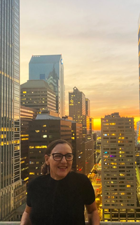 Julia Marsh, Artblog's new Executive Director and Editor, is wearing a black shirt and glasses stands in front of the sun setting on the Philadelphia skyline.