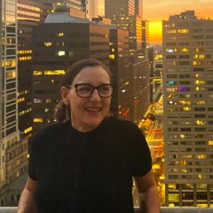 Julia Marsh, Artblog's new Executive Director and Editor, is wearing a black shirt and glasses stands in front of the sun setting on the Philadelphia skyline.