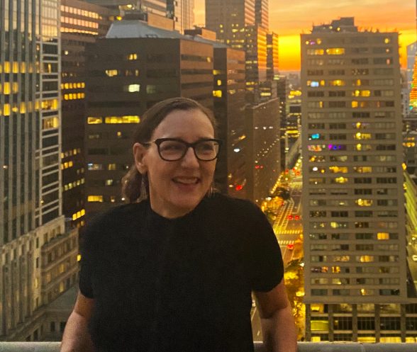 Julia Marsh, Artblog's new Executive Director and Editor, is wearing a black shirt and glasses stands in front of the sun setting on the Philadelphia skyline.