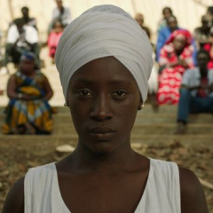In a still from the Senegalese film Xalé, a woman wearing a white head wrap and dress stares solemnly at the viewer while a large group of people who are out of focus stare at her from behind.