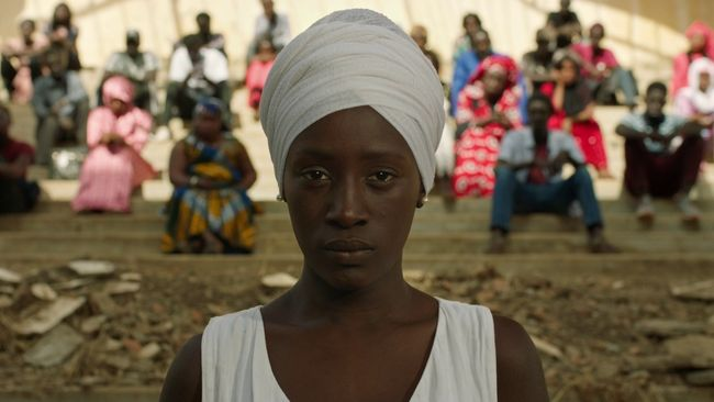 In a still from the Senegalese film Xalé, a woman wearing a white head wrap and dress stares solemnly at the viewer while a large group of people who are out of focus stare at her from behind.