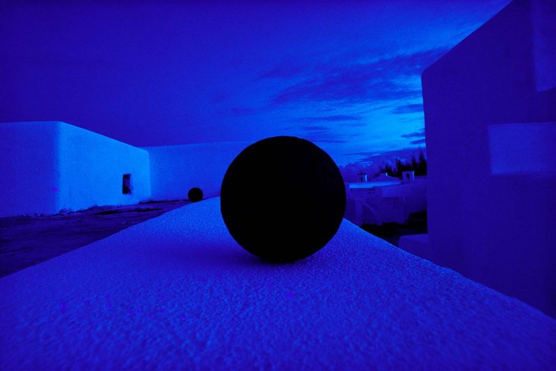A photo by the artist Pete Turner depicting a cannonball on top of a building surrounded by white walls that have been tinted blue.