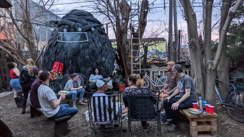 At the Open Kitchen Sculpture Garden, a group of people share a meal next to a large sculpture as the sun sets