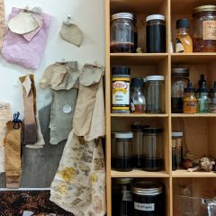 In an artist's studio, jars containing pigments and natural dyes are pictured next to fabric test samples hanging on the wall.