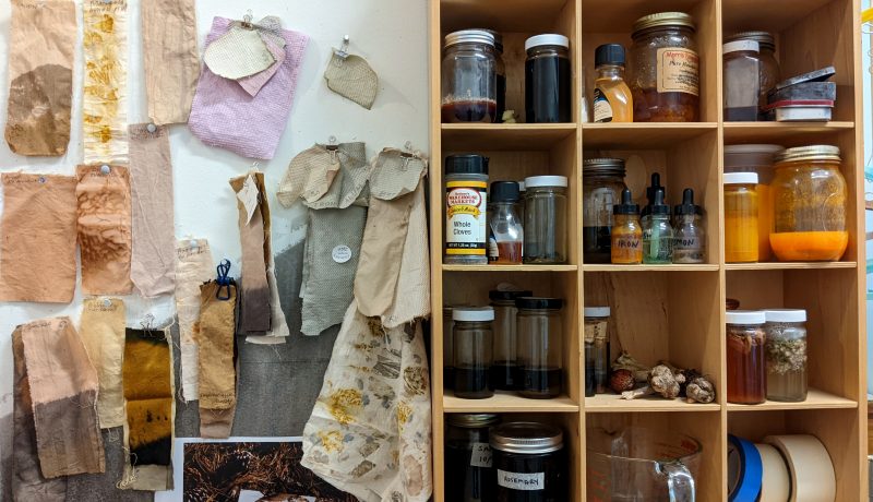 In an artist's studio, jars containing pigments and natural dyes are pictured next to fabric test samples hanging on the wall.