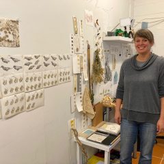 Artist Cindy Stockton more in her studio, smiling next to her nature-inspired artworks.