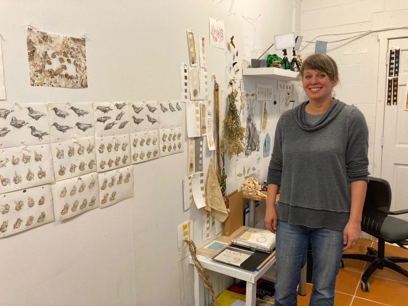 Artist Cindy Stockton more in her studio, smiling next to her nature-inspired artworks.
