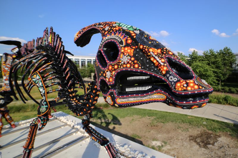 A color photo shows a colorful sculpture of a bead- and yarn-covered skeleton of a dinosaur in an outdoor space.