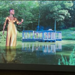 A color photo shows a beautiful lagoon surrounded by lush trees and brush, with a pavilion at the pond’s edge edge and a giant Black avatar wearing a gold dress standing in the water and gesticulating, while in the pavilion can be seen a human-sized woman in a silver-sparkly dress standing looking out at the avatar.