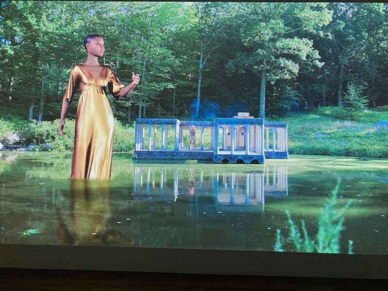 A color photo shows a beautiful lagoon surrounded by lush trees and brush, with a pavilion at the pond’s edge edge and a giant Black avatar wearing a gold dress standing in the water and gesticulating, while in the pavilion can be seen a human-sized woman in a silver-sparkly dress standing looking out at the avatar.