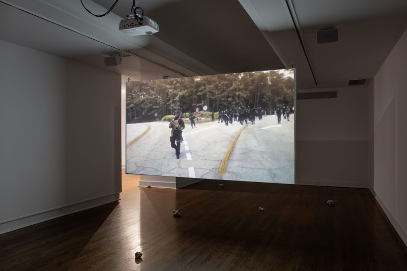 A color photo shows a dark room with a video projected onto a screen hanging in the space, and the video shows a group of black-clad, militia-types walking menacingly down an empty road towards the camera.